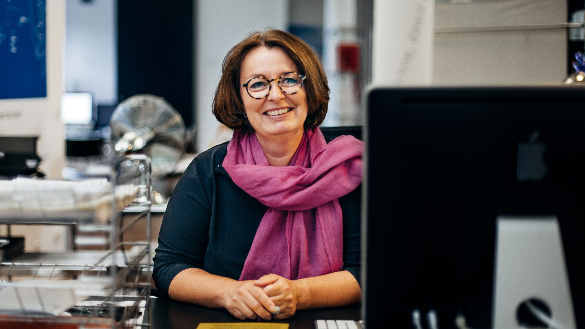 Mit einem großen violettem Schal sitzt Andrea lachend an ihrem Empfangstresen.