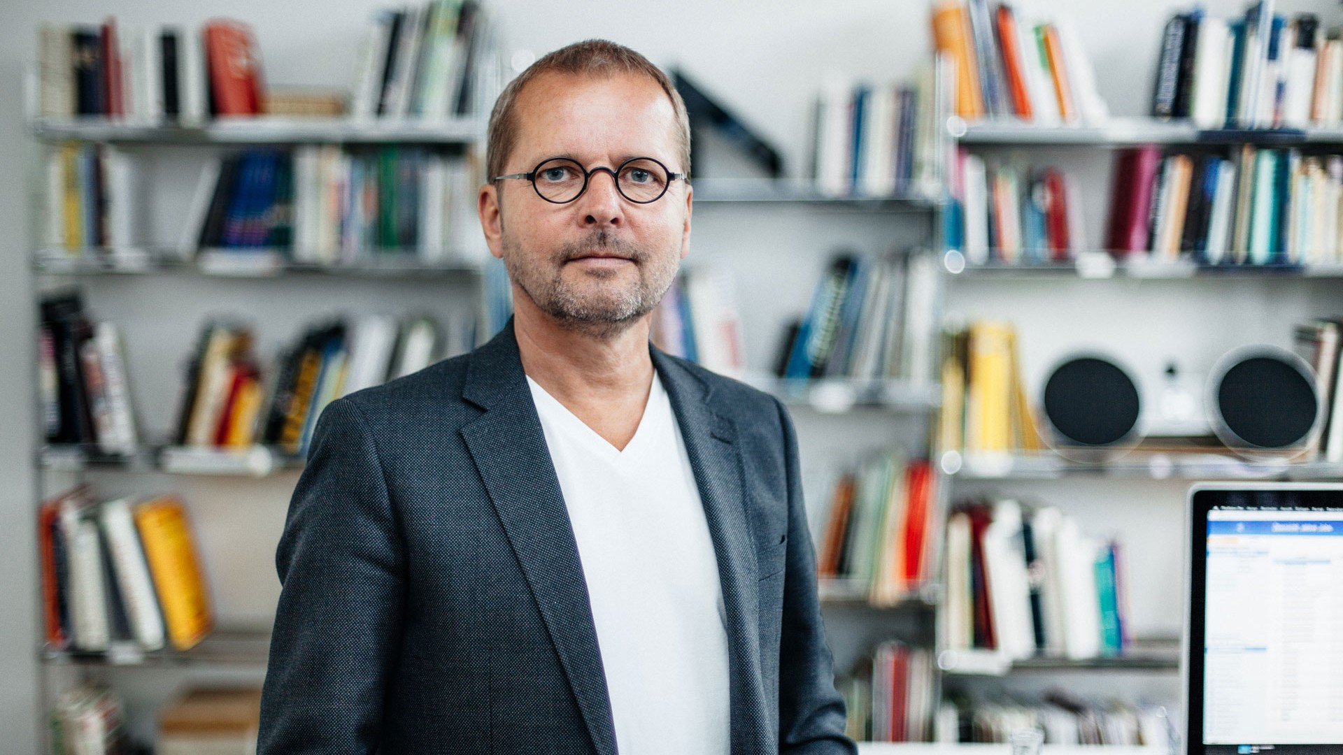 Florian steht in weißem T-Shirt und dunklem Sakko vor dem Bücherregal in seinem Büro. Er trägt eine Brille mit runden Gläsern und Dreitagebart.