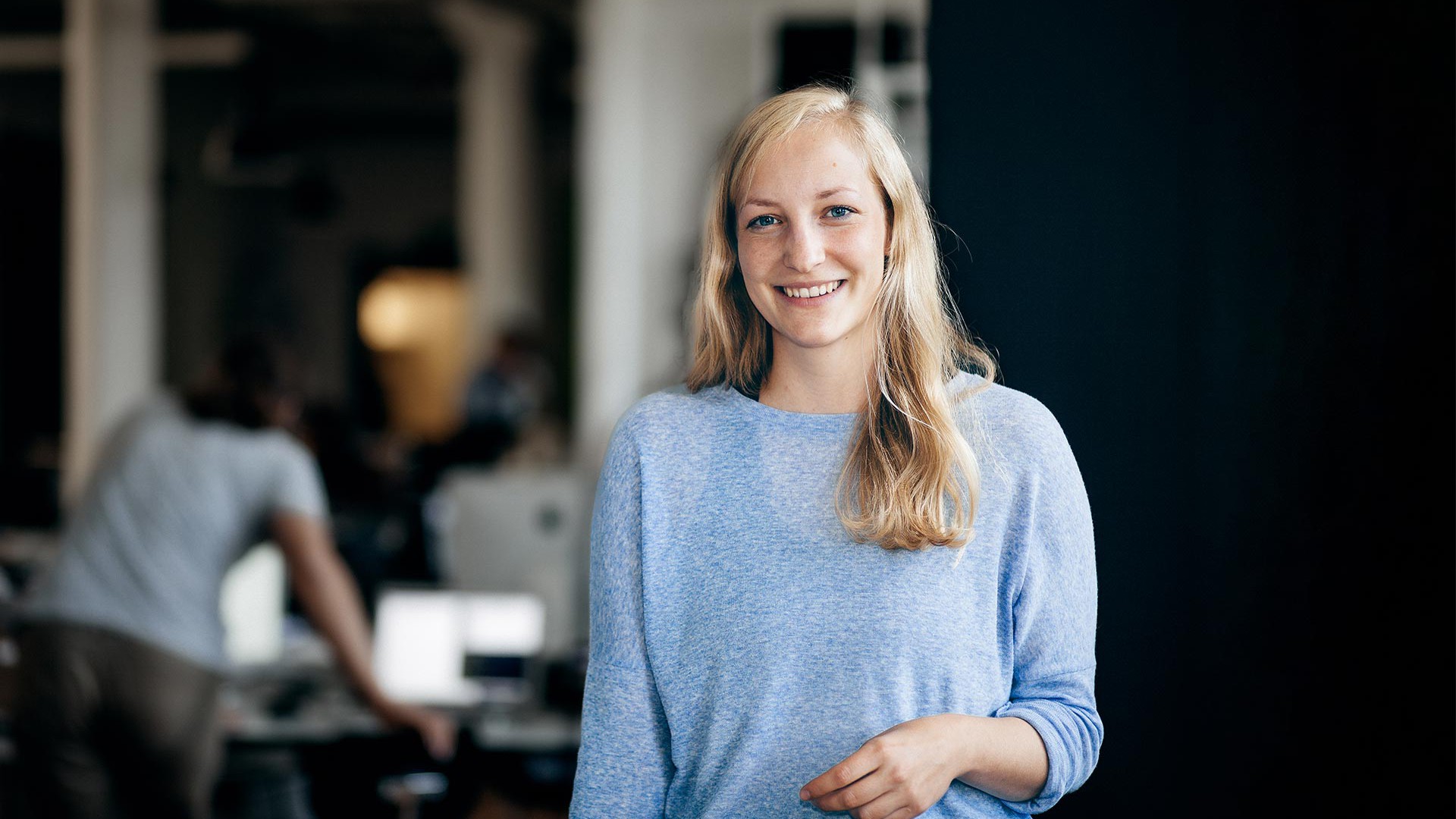 Johanna steht im Konferenzraum. Im Hintergrund arbeitet Björn an seinem Schreibtisch im Büro.