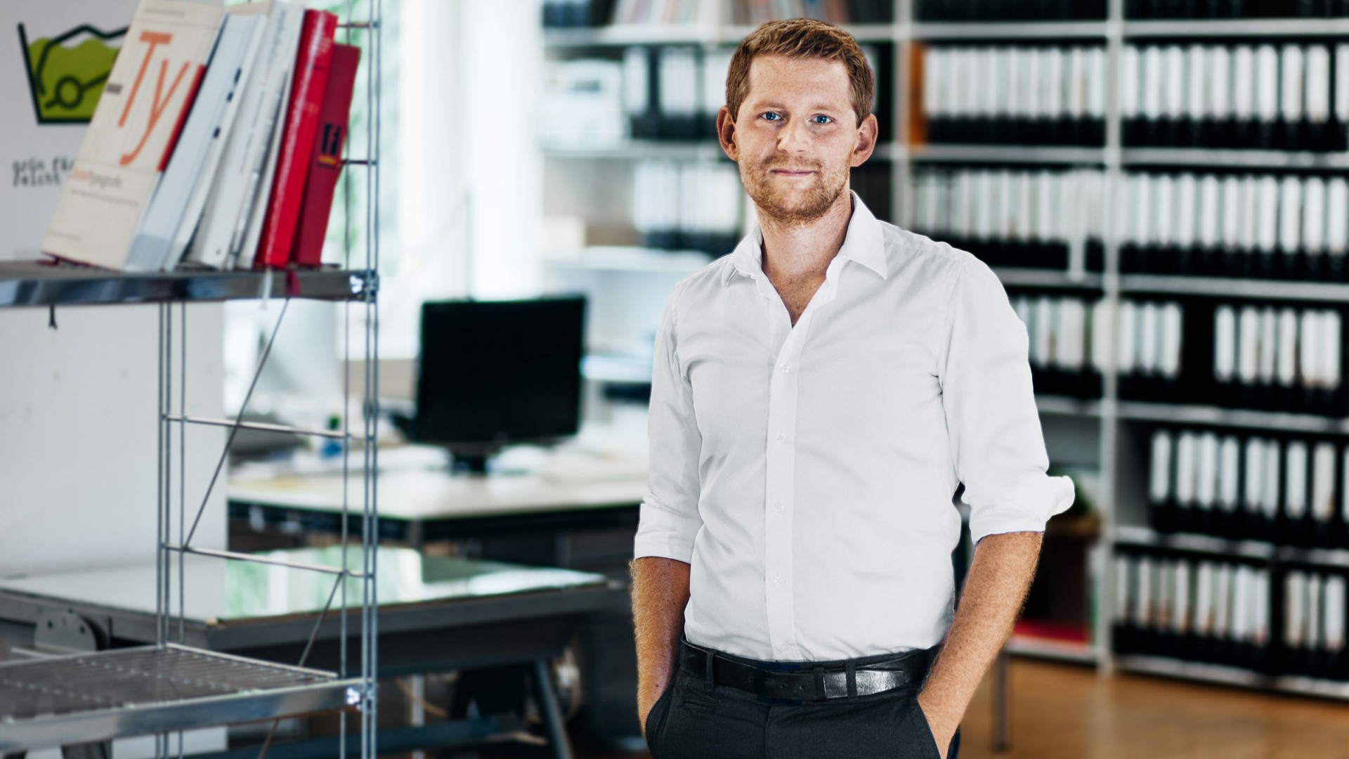 Tim steht in weißem Hemd zwischen Arbeitstischen und Bücherregalen im Büro.