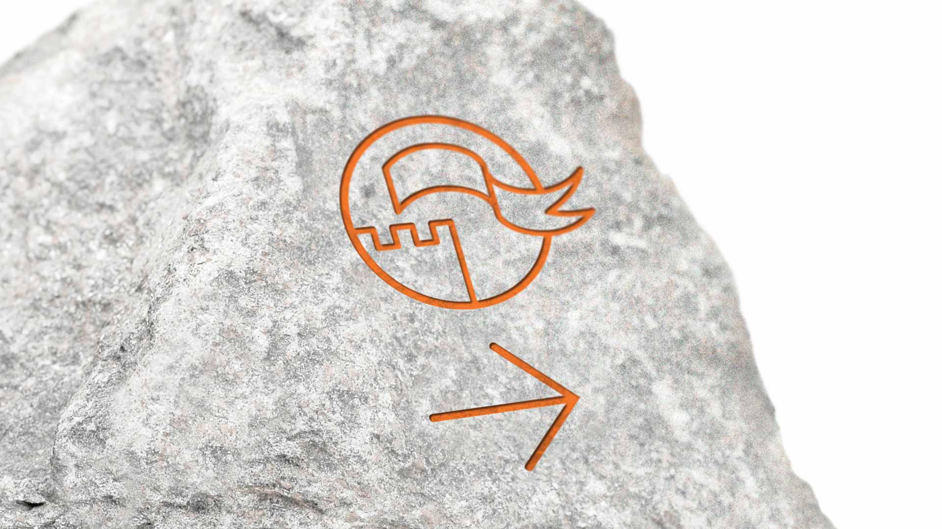 In einem Findling ist das Piktogramm für »Burgen« samt einem Richtungspfeil eingraviert und in orangener Farbe ausgelegt.